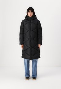 Long black puffer coat with a diamond quilted pattern, hood, and zipper pockets, paired with blue jeans and black shoes.