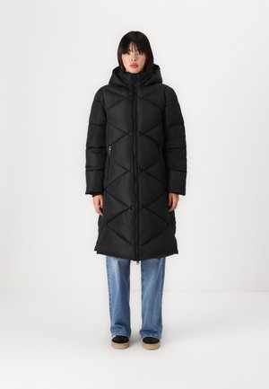 Longue doudoune noire avec un motif matelassé en losanges, capuche et poches zippées, associée à un jean bleu et des chaussures noires.