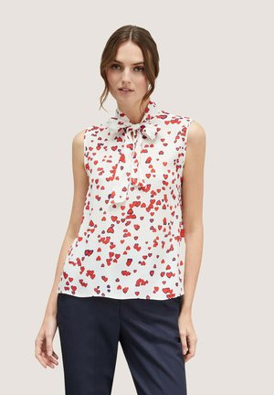 Mouwloze blouse van witte stof met een strik in de hals. Heeft een verspreid patroon van rode en paarse hartjes.
