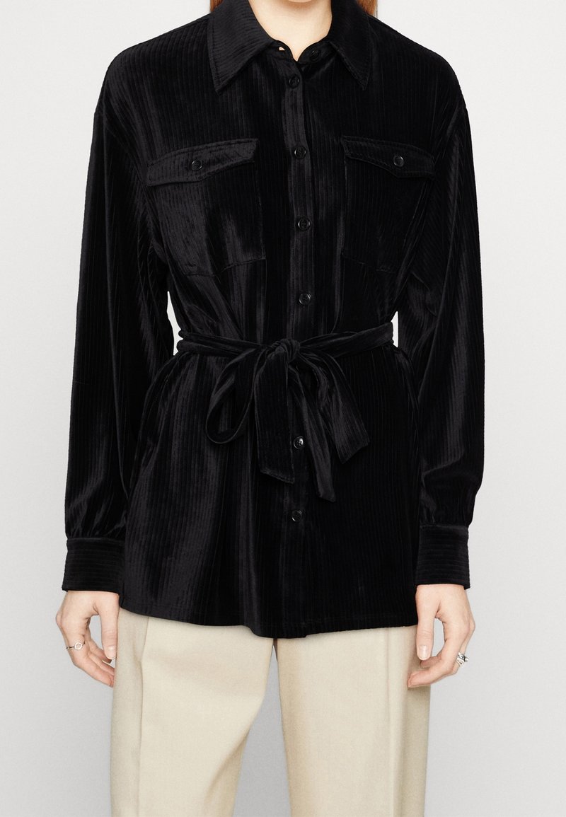 Chemise noire en velours côtelé à manches longues avec boutons sur le devant, poches poitrine et ceinture nouée à la taille, portée sur un pantalon beige.