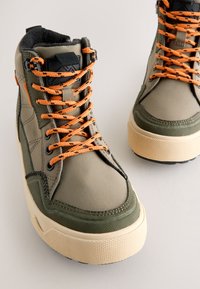 Baskets montantes en vert olive et beige. Fabriquées en tissu avec des lacets orange. Semelles texturées et col rembourré pour le confort.