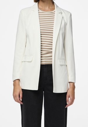 Femme portant un blazer blanc par-dessus un haut rayé beige et blanc avec un pantalon noir sur un fond blanc uni.