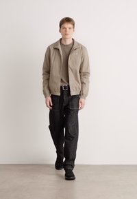 Giacca zip-up beige chiaro sopra un maglione khaki, abbinata a jeans scuri. Le scarpe nere completano l'outfit su uno sfondo neutro.