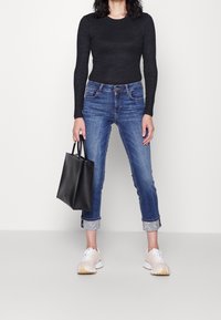 Top negro de manga larga, jeans de mezclilla azules con puños doblados que revelan un ribete estampado, bolso de mano negro y zapatillas beiges.