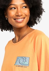 Orange t-shirt with a rounded neckline and short sleeves, featuring a blue graphic design with wave patterns and logo on the front.