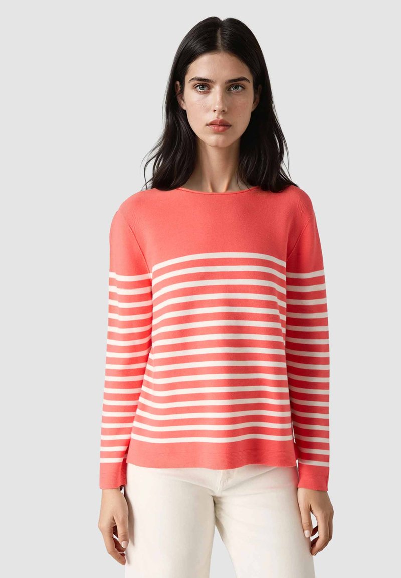 Kvinde med mørkt hår iført en koral- og hvidstribet langærmet sweater og hvide bukser, stående mod en ensfarvet lys baggrund.