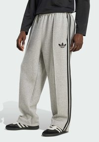 Sivé tepláky vyrobené z mäkkej tkaniny, s voľným strihom, čiernymi bočnými pruhmi a logom Adidas na ľavom stehne.