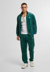 Chándal de terciopelo en verde oscuro con chaqueta de cremallera y pantalones a juego. Cuenta con un logo en el pecho y un bolsillo lateral, combinado con zapatillas blancas.