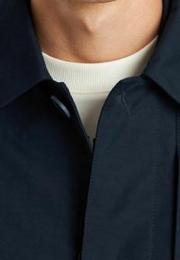 Primer plano del cuello de una persona que lleva una camiseta blanca de canalé bajo un abrigo oscuro de color marino con un botón y un cuello visibles.