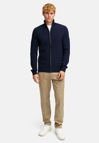 Granatowy, prążkowany kardigan zapinany na zamek wykonany z dzianiny, z wysokim kołnierzem i długimi rękawami, w połączeniu z beżowymi spodniami i białymi trampkami.