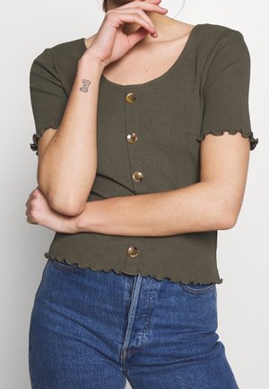 Olive green ribbed short-sleeve top with a scoop neckline, button closure, and ruffled edges. Paired with blue high-waisted jeans.