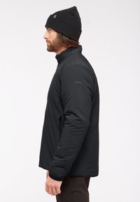 Zwart geïsoleerd jack met een hoge kraag, ritssluiting aan de voorkant en twee zijzakken. Effen zwarte beanie op het hoofd. Effen achtergrond.