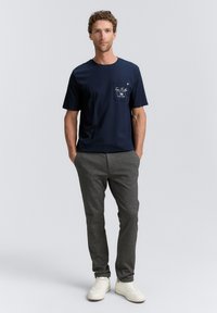 Mörkblå bomulls-T-shirt med tryckt fickdetalj. Matchad med grå smalnande byxor och vita sneakers. Enkel design och avslappnad passform.