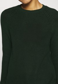 Maglione a maglia verde scuro con un motivo texturizzato, scollatura rotonda e maniche raglan. Il tessuto appare morbido e leggermente elasticizzato.