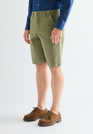 Mann trägt olivgrüne knielange Shorts, braune Schnürschuhe und ein langärmeliges blaues Hemd vor einem schlichten weißen Hintergrund.