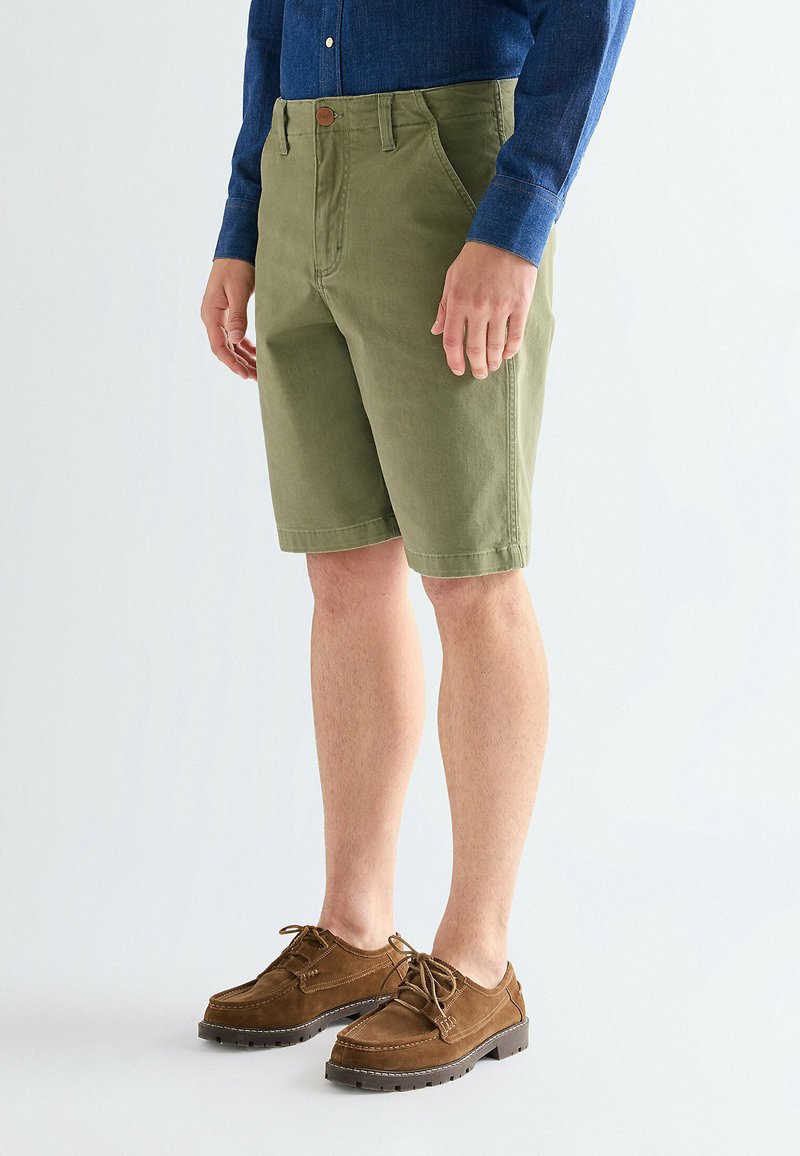 Homme portant un short olive longueur genou, des chaussures marron à lacets et une chemise bleue à manches longues sur un fond blanc uni.