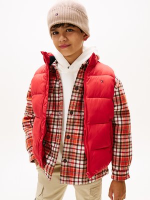 Gilet en duvet rouge sur une chemise en flanelle à carreaux avec des tons blancs, rouges et bruns, associé à un léger hoodie et un bonnet en tricot beige.