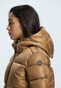 Doudoune kaki avec capuche, design matelassé et accent logo rond sur l'épaule. Tissu lisse et brillant avec des détails à cordon de serrage.