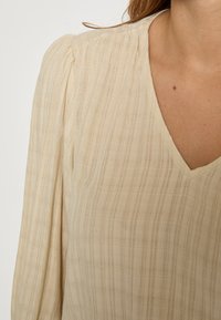 Béžová textúrovaná blúzka s dlhým rukávom a jemným károvaným vzorom a výstrihom do V, ktorú nosí osoba, vidieť hornú časť hrudníka a rameno.