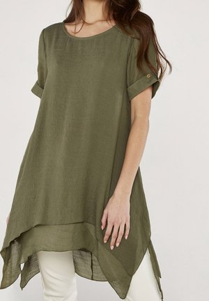 Kvinde iført en løs olivengrøn tunika med korte ærmer, asymmetrisk kant og knapdetalje på manchet, kombineret med hvide bukser.