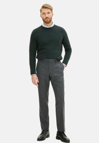 Dunkelgrüner Wollpullover über einem hellblauen Hemd mit Kragen, kombiniert mit grauen, maßgeschneiderten Hosen und schwarzen Leder-Schuhen. Gerade geschnitten, lässiger Stil.