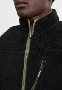 Veste polaire noire avec col montant, featuring des accents vert olive et des poches zippées. Détail de couture visible sur le tissu.