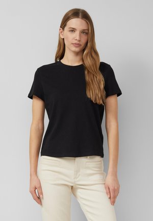 Frau mit langen hellbraunen Haaren, die ein schwarzes kurzärmeliges T-Shirt und hellbeige Hose trägt, steht vor einem einfarbigen grauen Hintergrund.