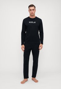 Schwarzes Langarmoberteil und passende Hose aus weichem Stoff. Das Oberteil hat den Text "REPLAY" in einer kontrastierenden weißen Schrift.