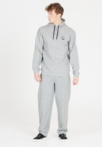 Grå hættetrøje med snore, side-logo, parret med matchende grå sweatpants. Modellen bærer sorte sneakers med hvid sål. Enkel, komfortabel design.