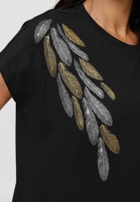 T-shirt noir avec broderie métallique de plumes en argent et or sur un côté. Manches courtes et design col rond.