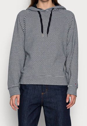 Persoon draagt een hoodie met zwart-wit pied-de-poule patroon, zwarte koordjes en donkerblauwe spijkerbroek, staand tegen een lichte achtergrond.