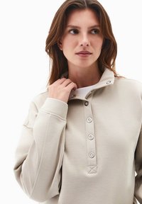 Jasno beżowy sweter z wysokim kołnierzem, dużymi guzikami i fakturowanym materiałem. Posiada wyhaftowane detale na kołnierzu i rękawach.
