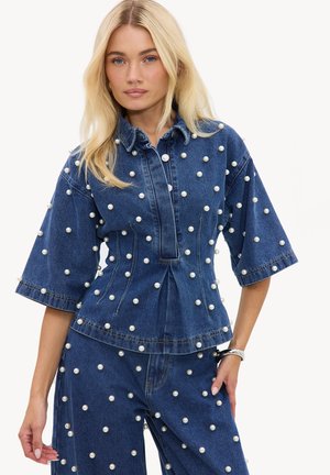 Femme blonde portant un haut en jean bleu et un pantalon, tous deux décorés de perles blanches espacées de manière uniforme.