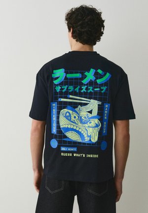 Person trägt ein schwarzes T-Shirt mit einem neonblauen und grünen Motiv einer Ramen-Schüssel mit Augen und Essstäbchen, der Text lautet "mysterious ramen soup".