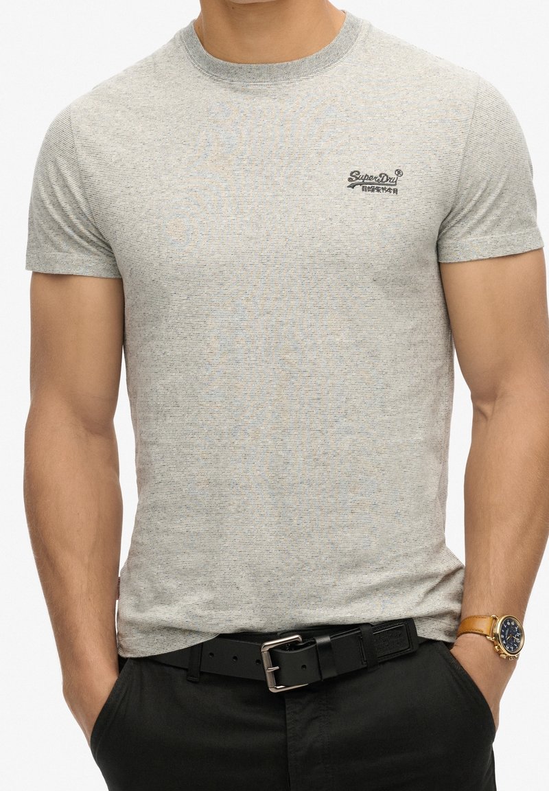 Homme portant un t-shirt Superdry gris à manches courtes, une ceinture noire, un pantalon noir et une montre-bracelet dorée avec un bracelet en cuir marron.