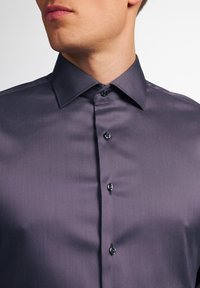 Męska koszula zapinana na guziki w ciemnym fiolecie, wykonana z gładkiej tkaniny, z klasycznym kołnierzykiem i czarnymi guzikami na przedniej listwie.