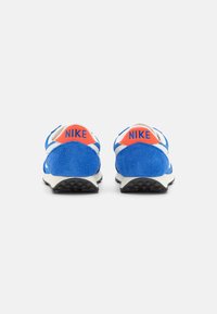 Blue suede sneakers with black outsole. Features white accents and orange Nike logo on the heel. Minimalist design with textured materials.