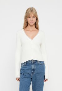 Νεαρή γυναίκα με ίσια ξανθά μαλλιά, ντυμένη με ένα λευκό ριμπ σακάκι V-λαιμό με δαντέλα και μπλε ψηλόμεσα τζιν, απέναντι από λευκό φόντο.