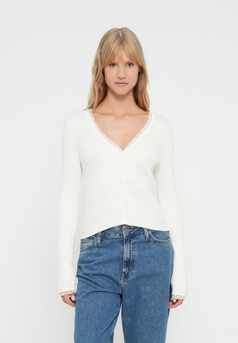 Νεαρή γυναίκα με ίσια ξανθά μαλλιά, ντυμένη με ένα λευκό ριμπ σακάκι V-λαιμό με δαντέλα και μπλε ψηλόμεσα τζιν, απέναντι από λευκό φόντο.