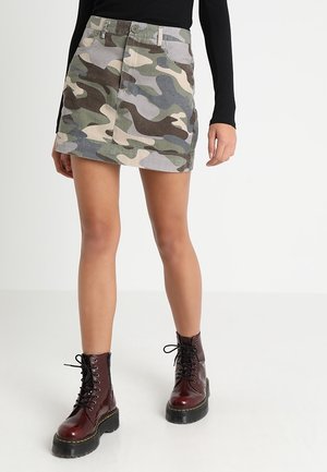 Femme portant une mini-jupe camouflage verte et marron, un haut noir à manches longues et des bottines à plateforme marron foncé avec des lacets.