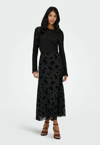 Donna con lunghi capelli scuri che indossa un top nero a maniche lunghe texturizzato e una gonna lunga nera con motivo floreale, insieme a dei tacchi neri con cinturini.