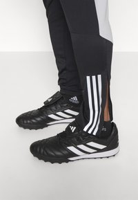 Schwarze Fußballschuhe mit weißen Akzenten, mit einer strukturierten Oberfläche und drei weißen Streifen. Schwarze Sporthosen mit grauen Seitenpaneelen.