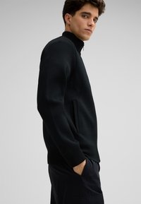 Zwarte textuurtrui met een hoge kraag, zijrits, geribde manchetten en een voorzak. Draagt met donkere broek, met een ontspannen pasvorm.