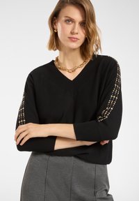 Czarny sweter V-neck z rękawami o długości 3/4, zdobiony dekoracyjnymi koralikami na rękawach. Noszony z szarymi spodniami.