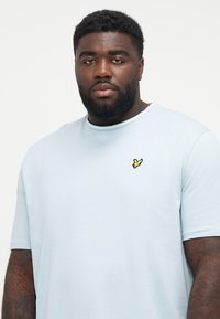 Lyseblå T-shirt med korte ærmer lavet af blød bomuld, med et lille gult broderet logo på brystet og rund halsudskæring.