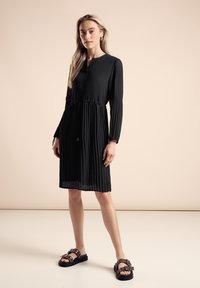 Robe noire plissée avec des manches longues, boutons au col et taille cintrée. Assortie avec des sandales noires avec des éléments décoratifs.