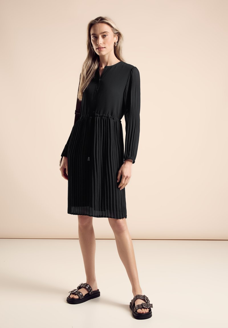 Robe noire plissée avec des manches longues, boutons au col et taille cintrée. Assortie avec des sandales noires avec des éléments décoratifs.