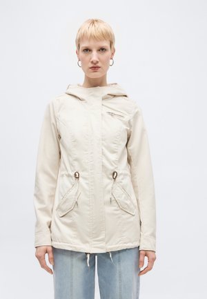 Kvinne iført en kremfarget hettejakke med glidelåslommer og dragsnorer, kombinert med lyseblå jeans, stående mot en ensfarget bakgrunn.