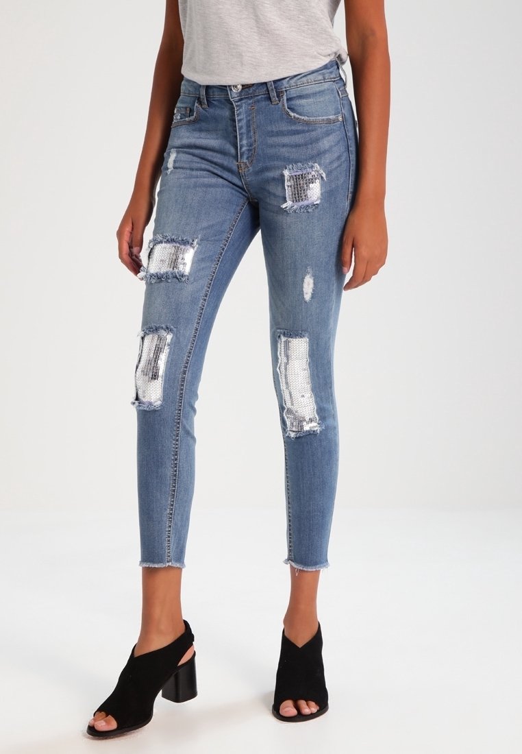 Jeans ajustados en denim azul de lavado medio con múltiples parches desgastados y dobladillos deshilachados. Combinados con tacones negros de punta abierta.