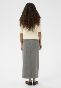 Kvinde med langt krøllet hår iført en cremefarvet ribbet top, grå ankel-længde nederdel og sorte balletflade sko, der står foran en enkel hvid baggrund.